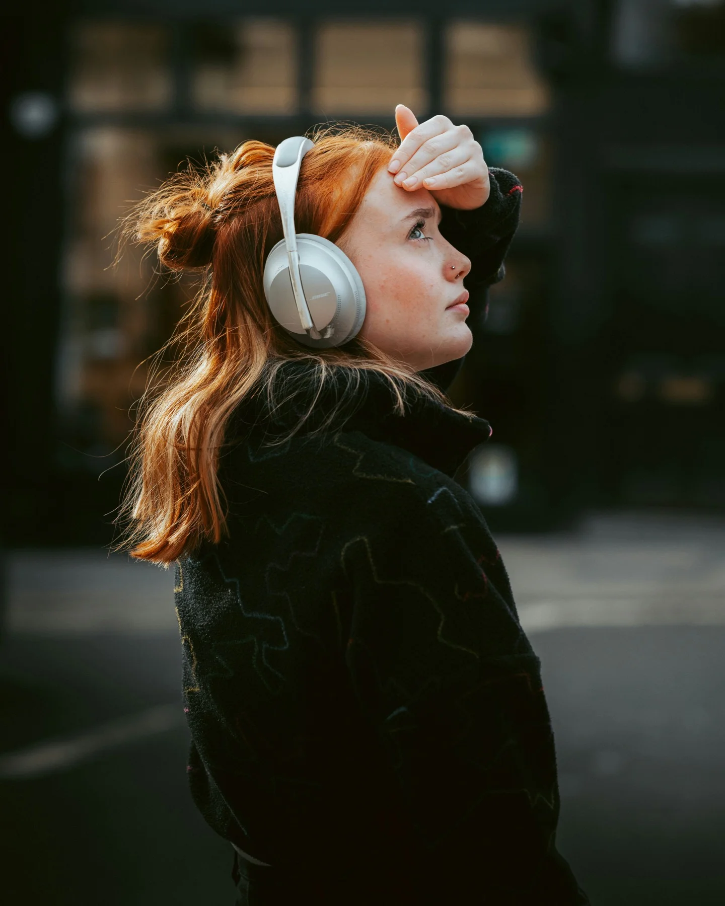A person wearing over-ear headphones on a city street, shading their eyes and looking up.
